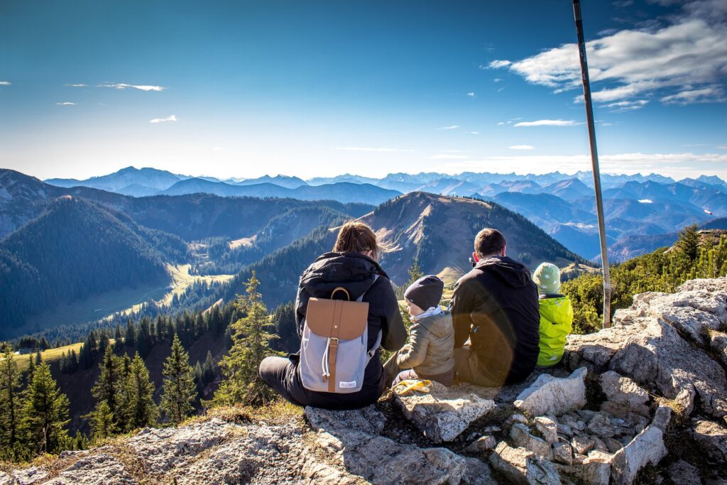 Família junta senta sobre pedras de uma montanha olhando para o horizonte, vendo montanhas e florestas à frente com um céu azul, representa ideia de atividade ao ar livre para o Dia dos Pais.