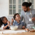 Pai na cozinha com duas crianças, uma menina e um menino, ilustra ideias de atividades para o Dia dos Pais 2024.